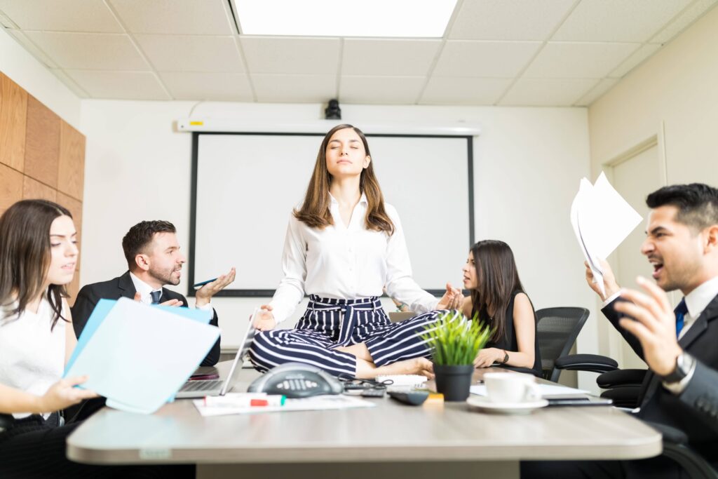 A woman meditating at her desk while colleagues around her engage in stressed and conflict-ridden interactions, illustrating how we can control how contagious we are despite our surrounding circumstances.